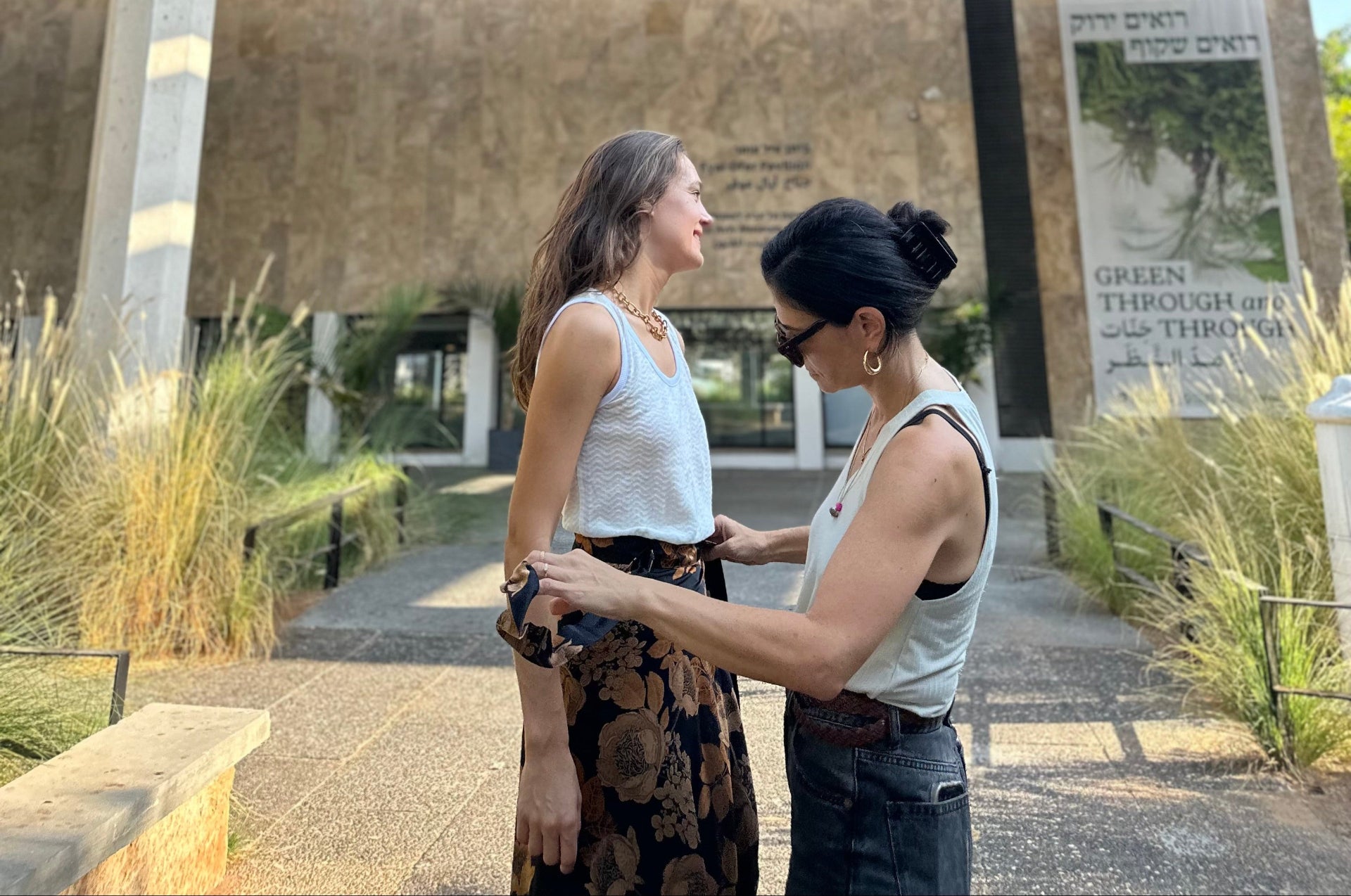 Two women standing outdoors, one adjusting the other's sleeve, with a building and sign in the background.